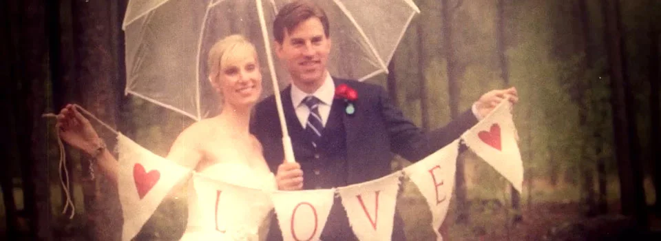 Bride and groom under a clear umbrella holding a LOVE banner in the woods